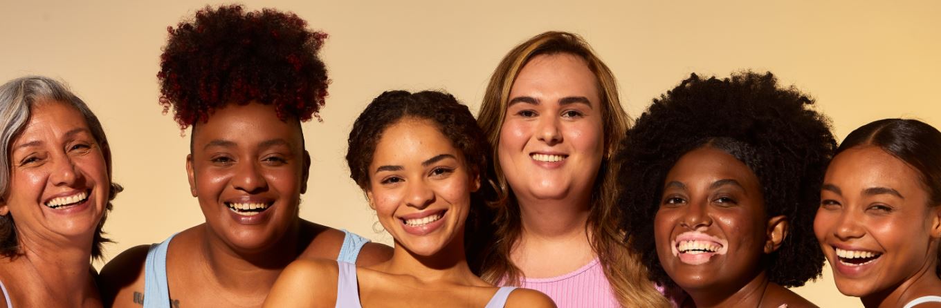 Foto de seis mulheres de variadas idades sorrindo olhando para câmera. Atrás delas tem um fundo laranja