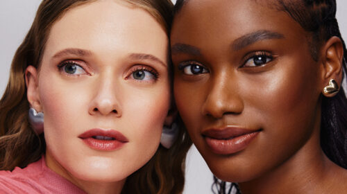 Fotografia em plano fechado de duas mulheres com tons de pele diferentes posicionadas lado a lado, destacando uma maquiagem iluminada e natural. Elas usam brincos em formato de coração.