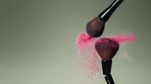 A imagem mostra dois pincéis da linha MakeB depositando uma névoa de blush rosa sobre um plano de fundo neutro na cor marrom.