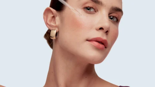 Fotografia de close-up do rosto de uma mulher jovem, com maquiagem natural, aplicando um sérum com um conta-gotas na bochecha. Ela usa um brinco de argola dourado e tem o cabelo preso.